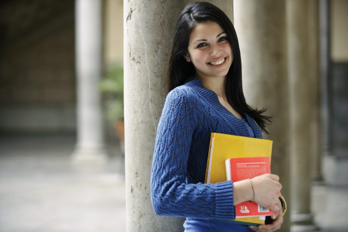 Young woman leaning against column holding books, smiling, portrait